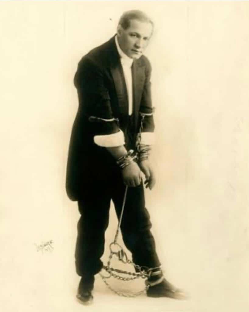 A man in a dark suit stands with his wrists, arms, and ankles bound in chains and handcuffs, looking slightly downward.
